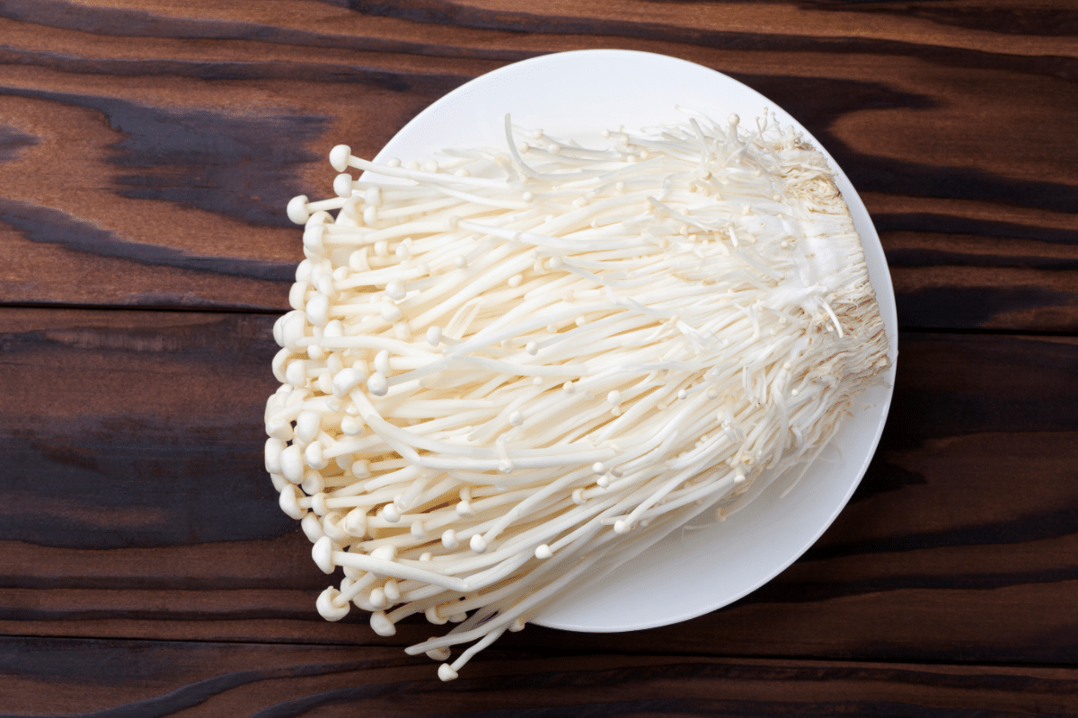 A plate of fresh enoki mushrooms with long, thin white stems and small caps, placed on a wooden surface.