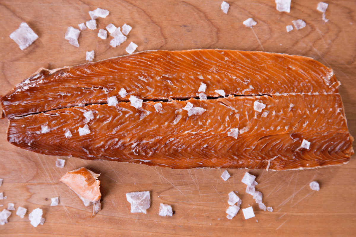 Close-up of a piece of smoked salmon fillet with a rich orange color and flaky texture, sprinkled with coarse sea salt on a wooden cutting board.