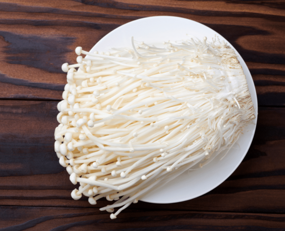 A plate of fresh enoki mushrooms with long, thin white stems and small caps, placed on a wooden surface.